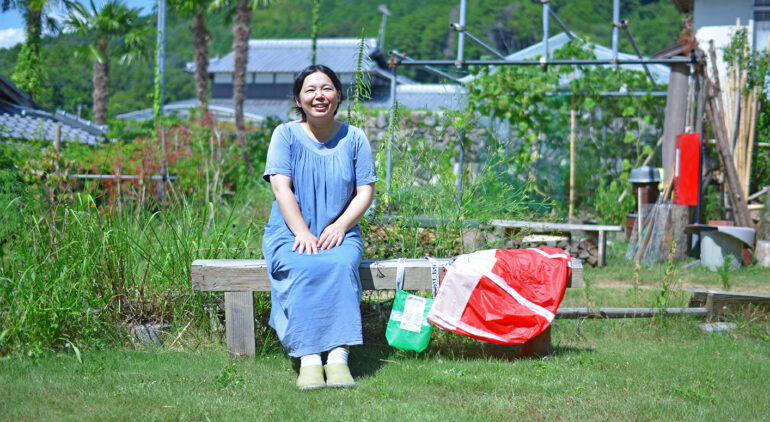 のどかな緑の芝生が広がる庭で、HOZUBAGを持ってベンチに座る工場長の武田さん。