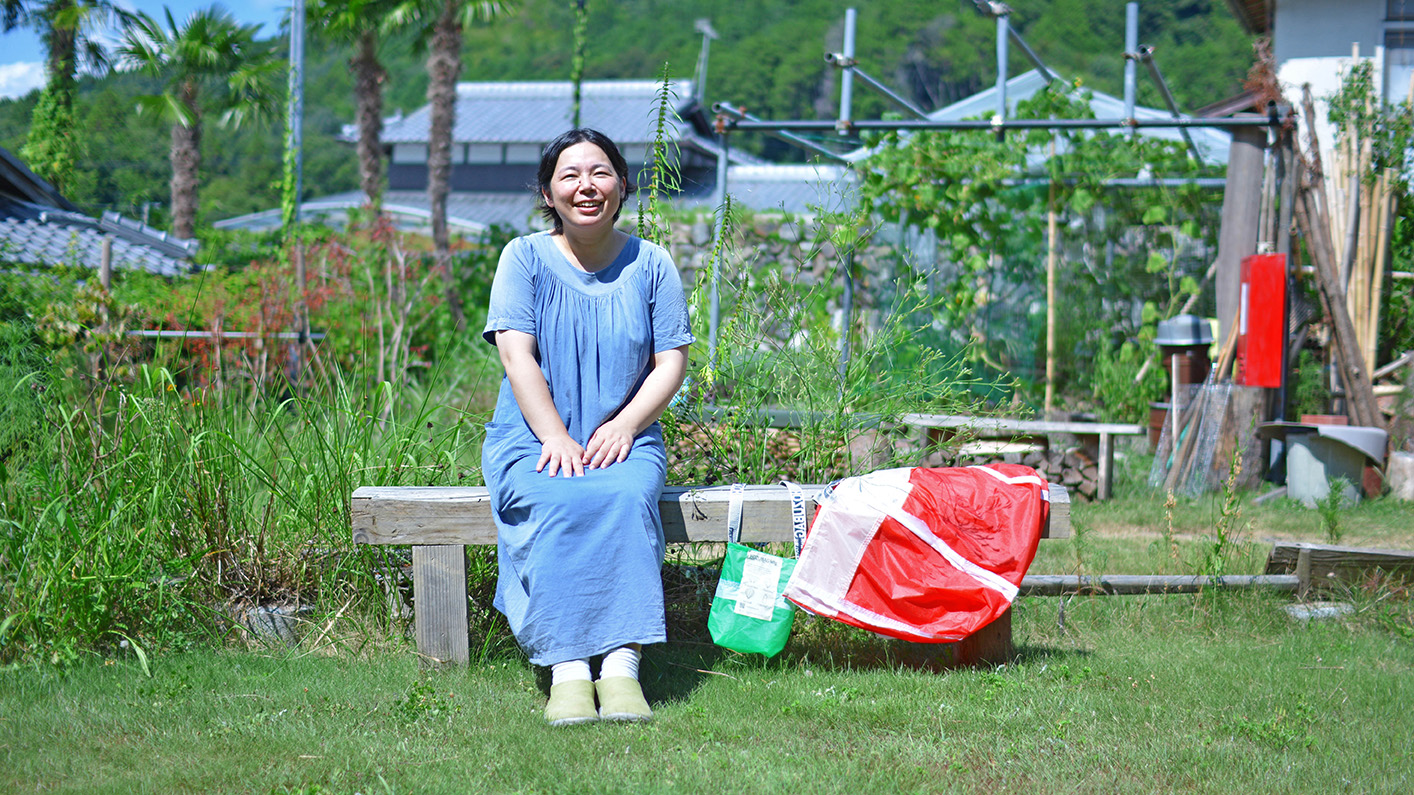 のどかな緑の芝生が広がる庭で、HOZUBAGを持ってベンチに座る工場長の武田さん。